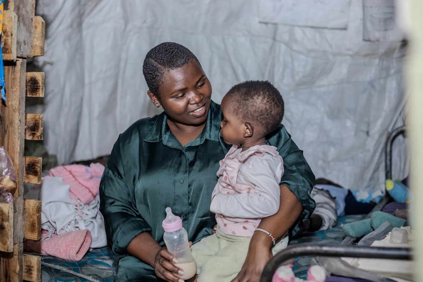 A daycare caregiver smiles at a baby on her lap, holding the baby's bottle of milk