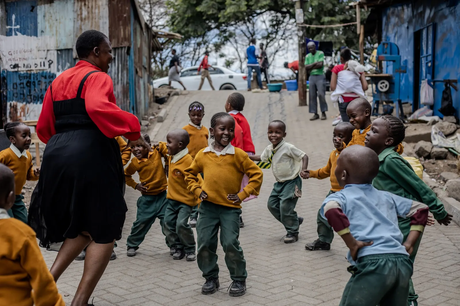 A teacher leads children in a circle game in the lane outside school