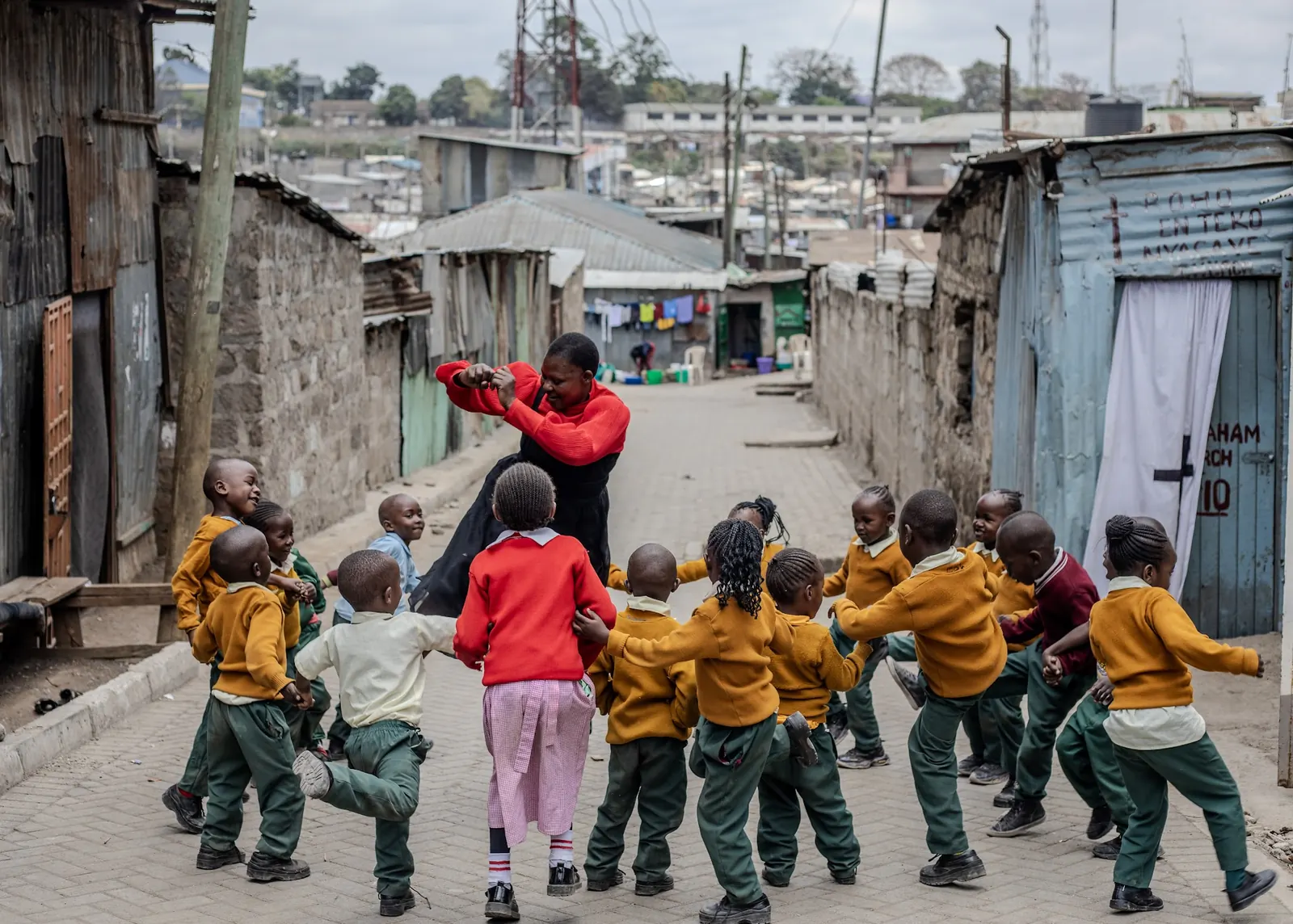 Children playing a circle game in the lane with a teacher, the rooftops of Mathare behind them