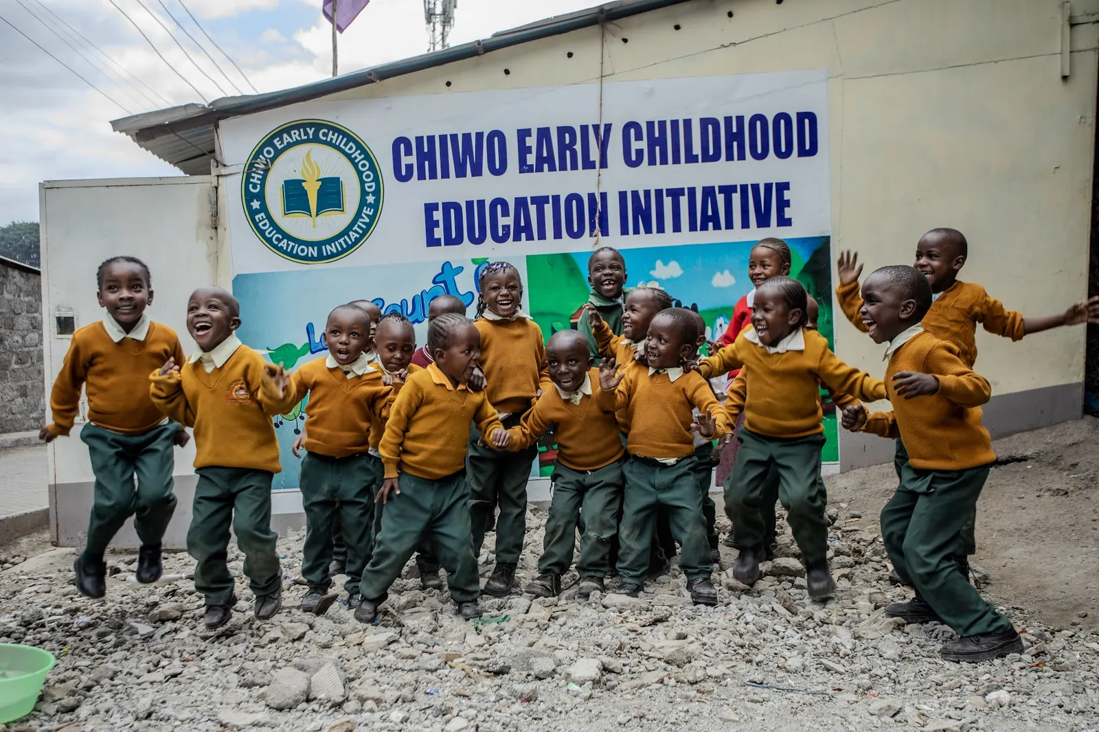 Children in their golden-yellow CHIWO uniforms jumping for joy in front of the school's painted sign