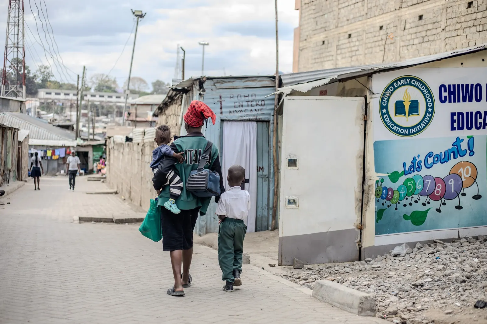 A mother carrying a baby walks a young pupil to school past the CHIWO sign